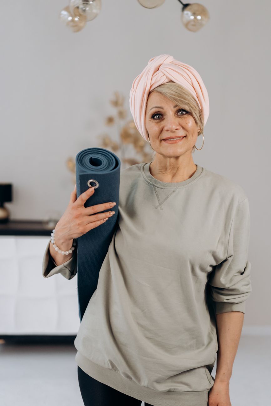 Femme souriante aux cheveux couverts d’un foulard rose, un tapis de yoga roulé sous le bras