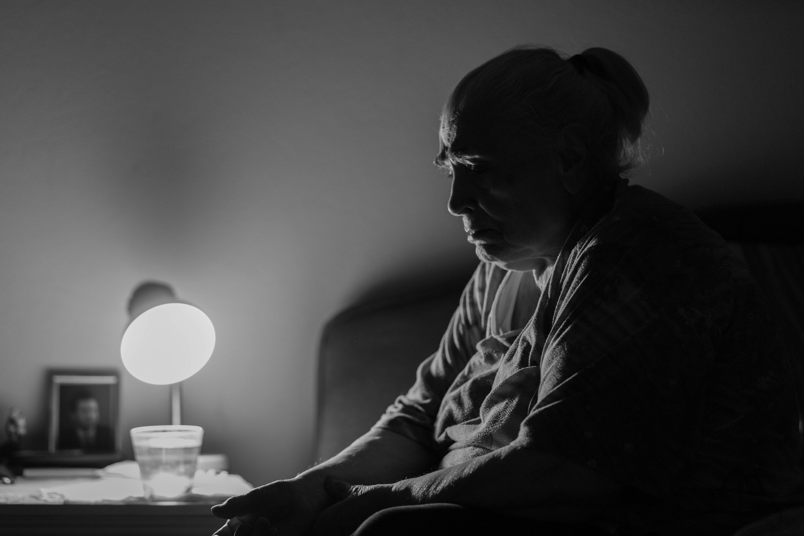 A contemplative senior woman sits by a lamp in a dimly lit room, evoking deep thought and solitude. Une femme âgée contemplative assise près d'une lampe dans une pièce faiblement éclairée, évoquant une profonde réflexion et la solitude.