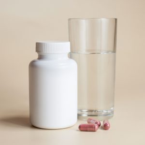 White supplement bottle with capsules and a glass of water on a neutral background. Bouteille de supplément blanc avec des capsules et un verre d'eau sur un fond neutre.