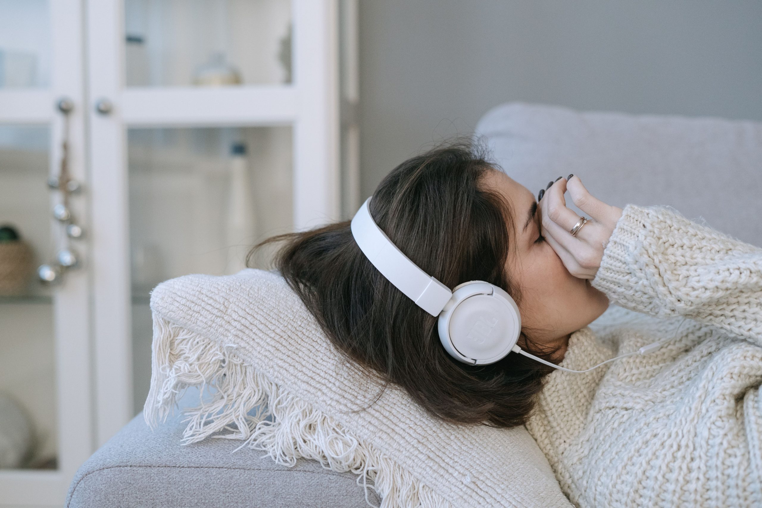 Woman lying on couch with headphones, appearing relaxed or contemplative indoors, cozy atmosphere. Femme allongée sur un canapé avec des écouteurs, l'air détendue ou contemplative à l'intérieur, atmosphère chaleureuse.