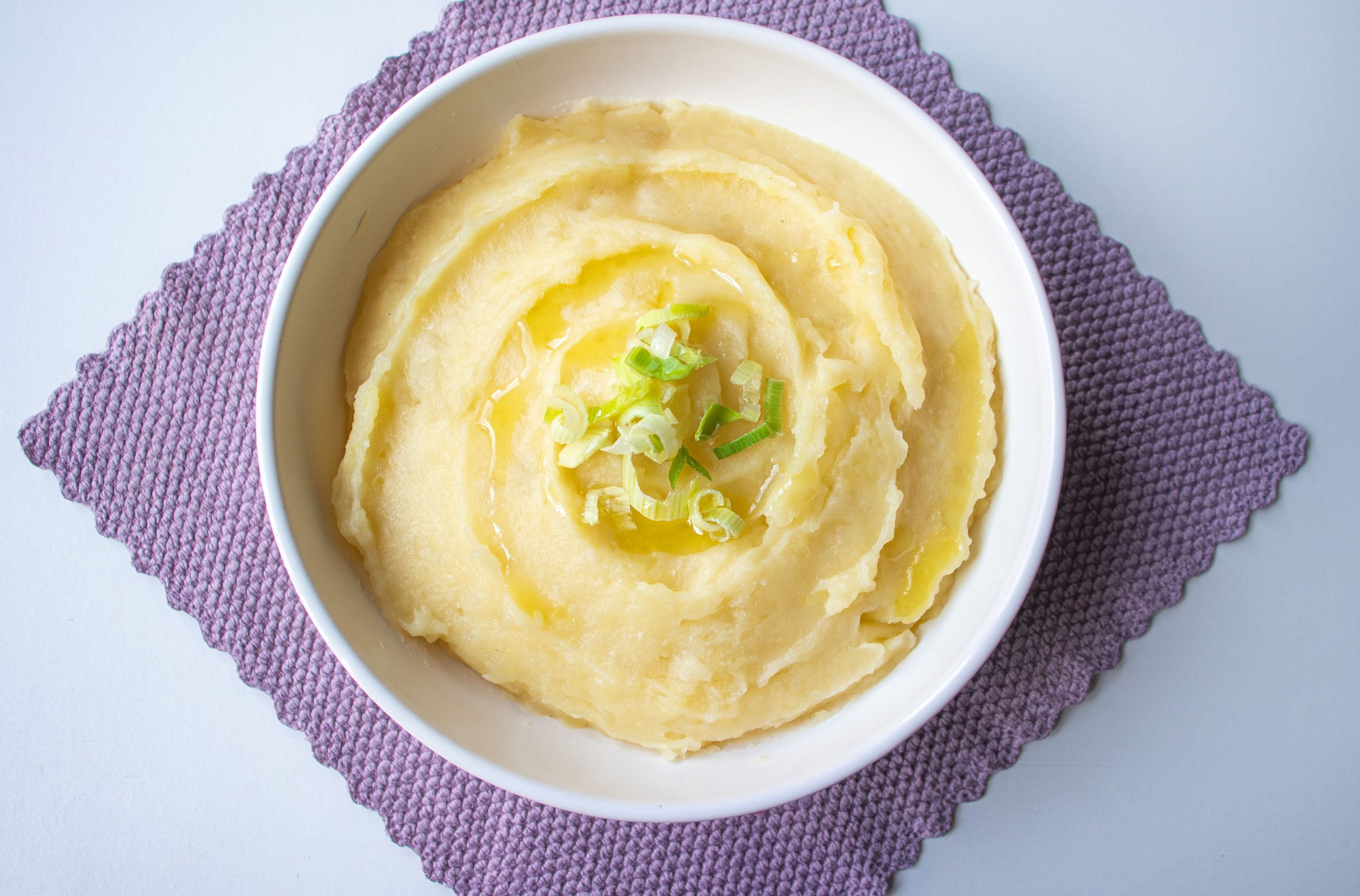 Top-down view of creamy mashed potatoes garnished with scallion in a white bowl. Vue de dessus de purée de pommes de terre crémeuse garnie d'oignon vert dans un bol blanc.