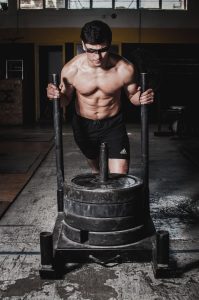 Strong shirtless man pushing a weighted sled indoors in a gym setting. Homme musclé torse nu poussant un traîneau lesté à l'intérieur d'une salle de sport.