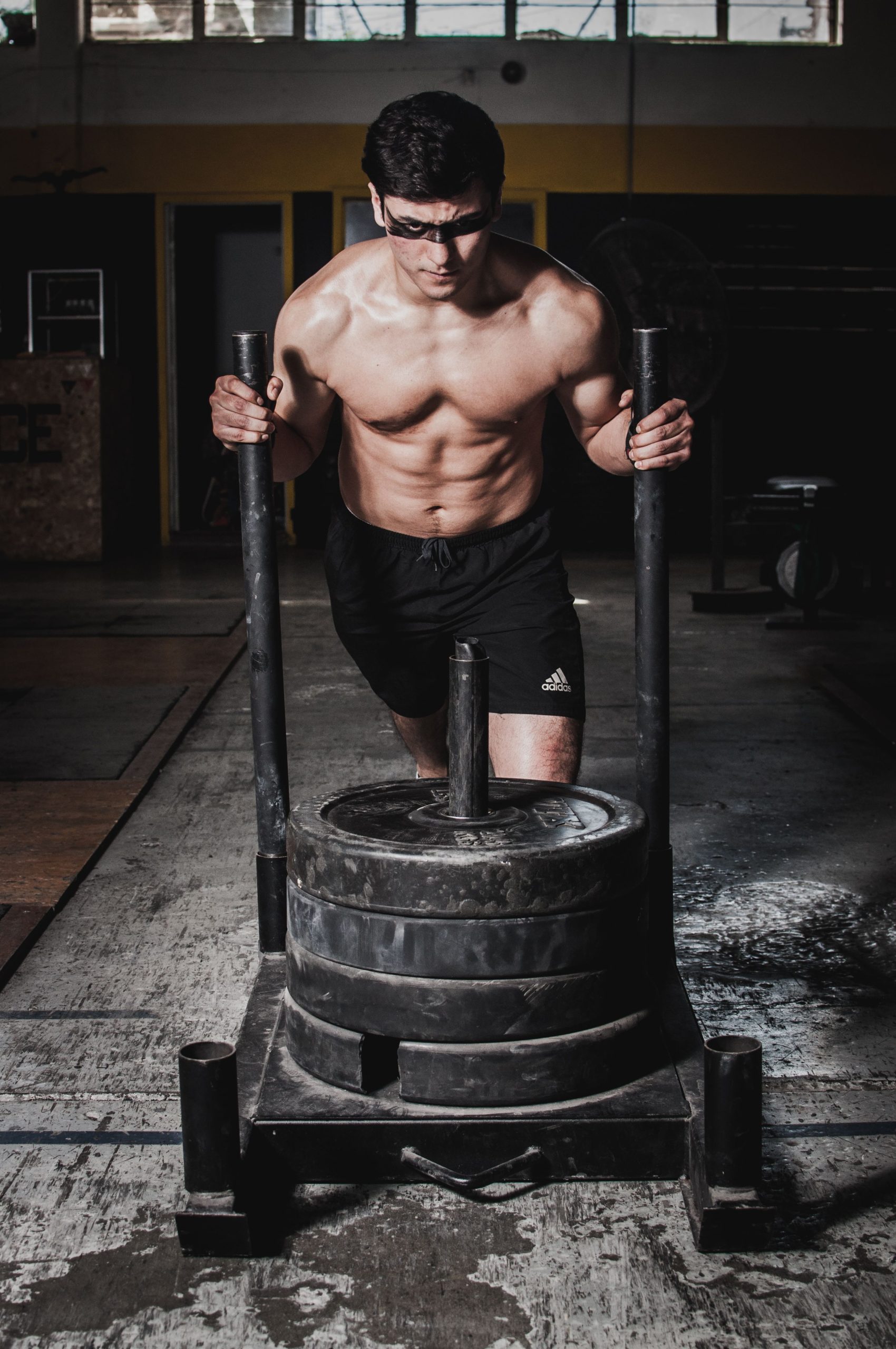 Strong shirtless man pushing a weighted sled indoors in a gym setting. Homme musclé torse nu poussant un traîneau lesté à l'intérieur d'une salle de sport.
