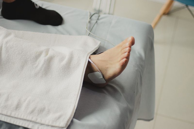 Électrode d’électrostimulation posée sur la cheville d’une personne allongée sur une table de soin, jambe sous une serviette